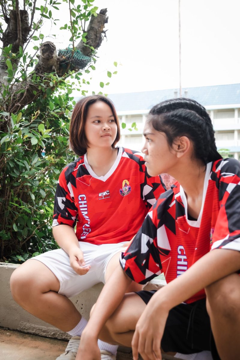 Two teenage girls in red soccer jerseys sitting outdoors, engaged in a conversation.